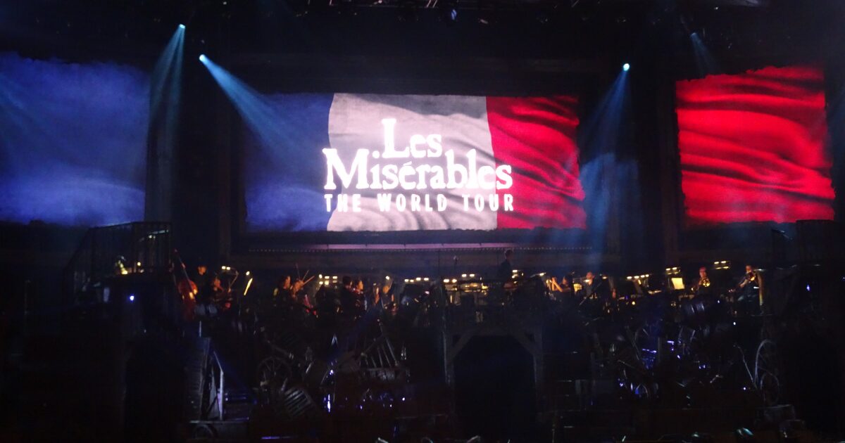2025年9月大阪上演の「レ・ミゼラブルワールドツアースペクタキュラー」二幕開演前のステージ（フランス国旗にレミゼラブルと書かれた巨大モニター映像の前にオーケストラが待機している）