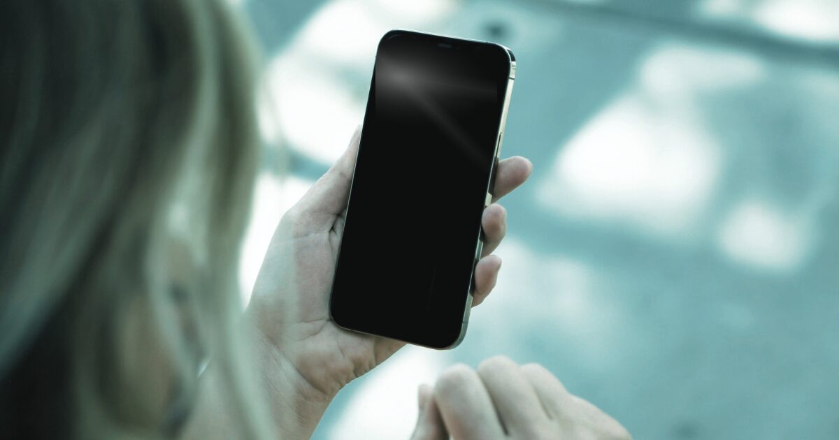 少し白く光った黒いスマホ画面を見つめる女性を背後から撮影した写真