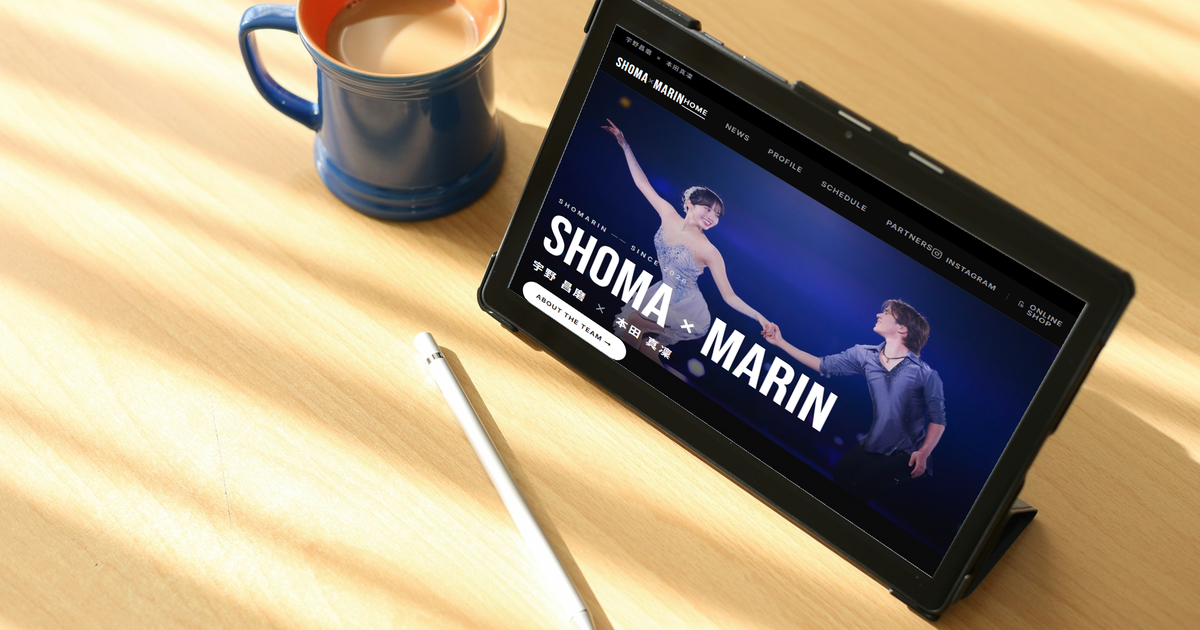 木のテーブル上にコーヒーカップとペンと「しょまりん」公式サイトが映っているタブレットが並んでいる写真　